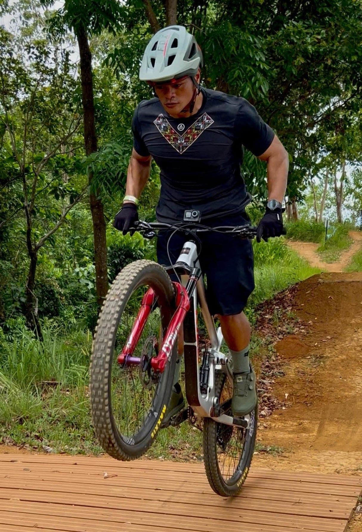 Male mountain biker jumping the bike while wearing the Uleyan Super Light SS Shirt in dark gray with colorful woven-inspired chest pattern