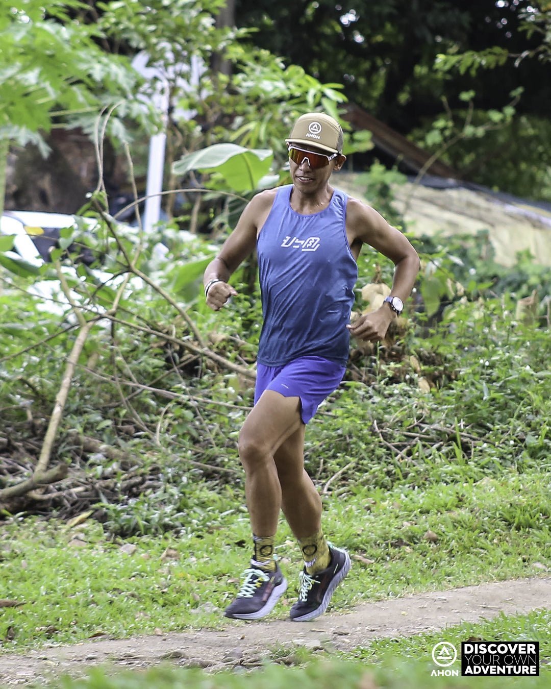 trail runner in Ahon blue green Ultra Light Singlet running through forest trail, wearing a cap and sunglasses