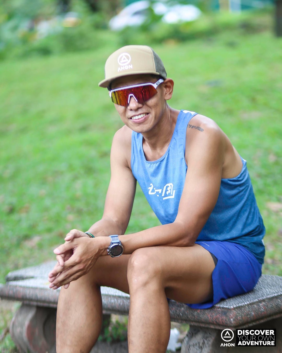 Male trail runner in Ahon blue green Ultra Light Singlet running through forest trail, wearing a cap and sunglasses